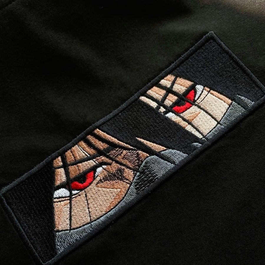 Close view of a rectangular embroidered panel patch on black fabric, showing a pair of eyes in dense satin stitch