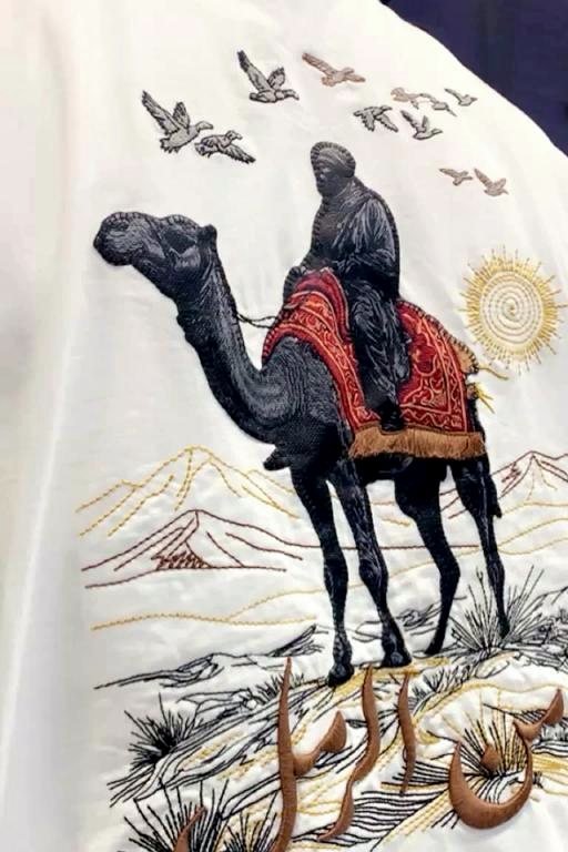 Bone-coloured oversized t-shirt, back view, embroidered with a robed rider on a camel crossing dunes under a gold sun, with Arabic script worked into the sand